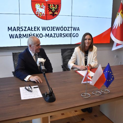 kobieta i mężczyzna siedzą, przed nimi dokumenty, w tle flagi Warmii i Mazur, Polski i Unii Europejskiej