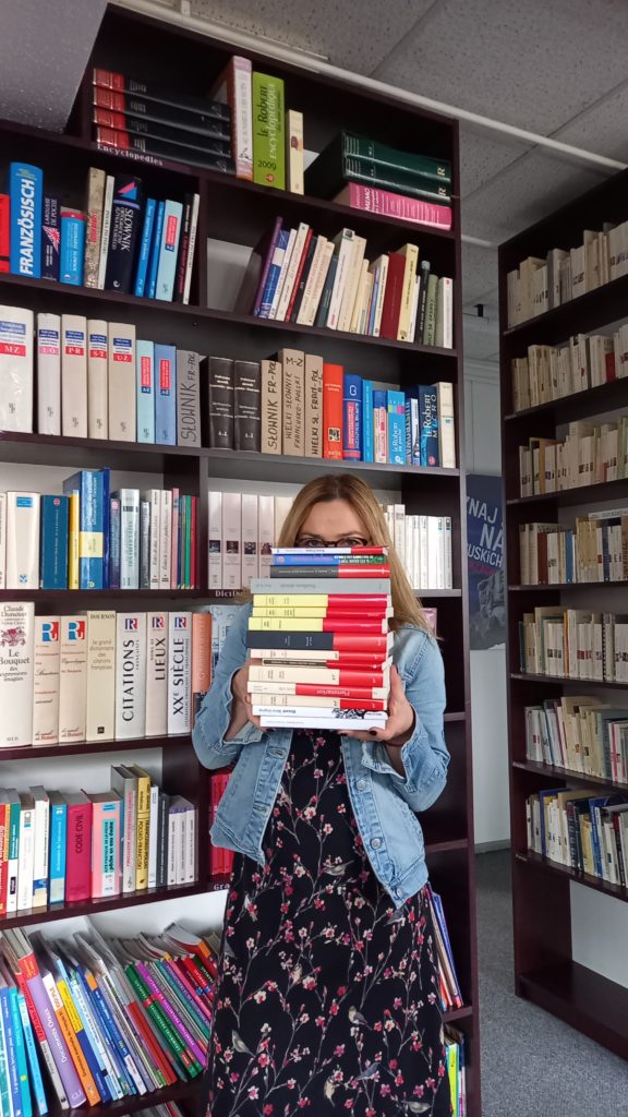 kobieta trzyma przed sobą stos książek, znajduje się w bibliotece, pełnym regałów