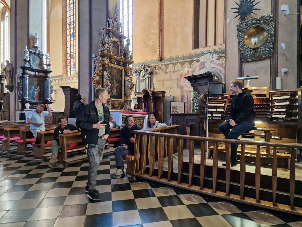 Grupa kilku osób zgromadzona w kościele, mężczyzna siedzący przed organami opowiada 
